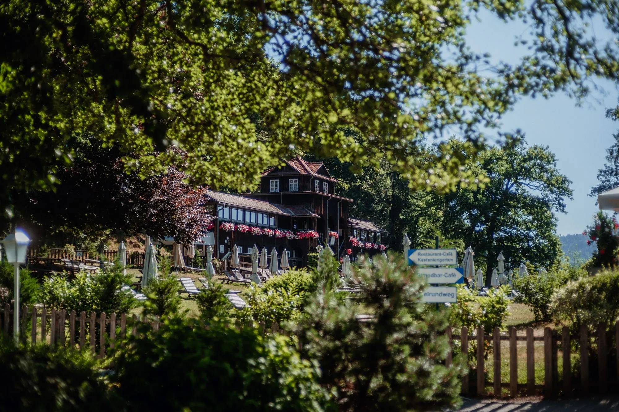 Das Badehaus zwischen den Bäumen, davor der Zaun und der Weg.