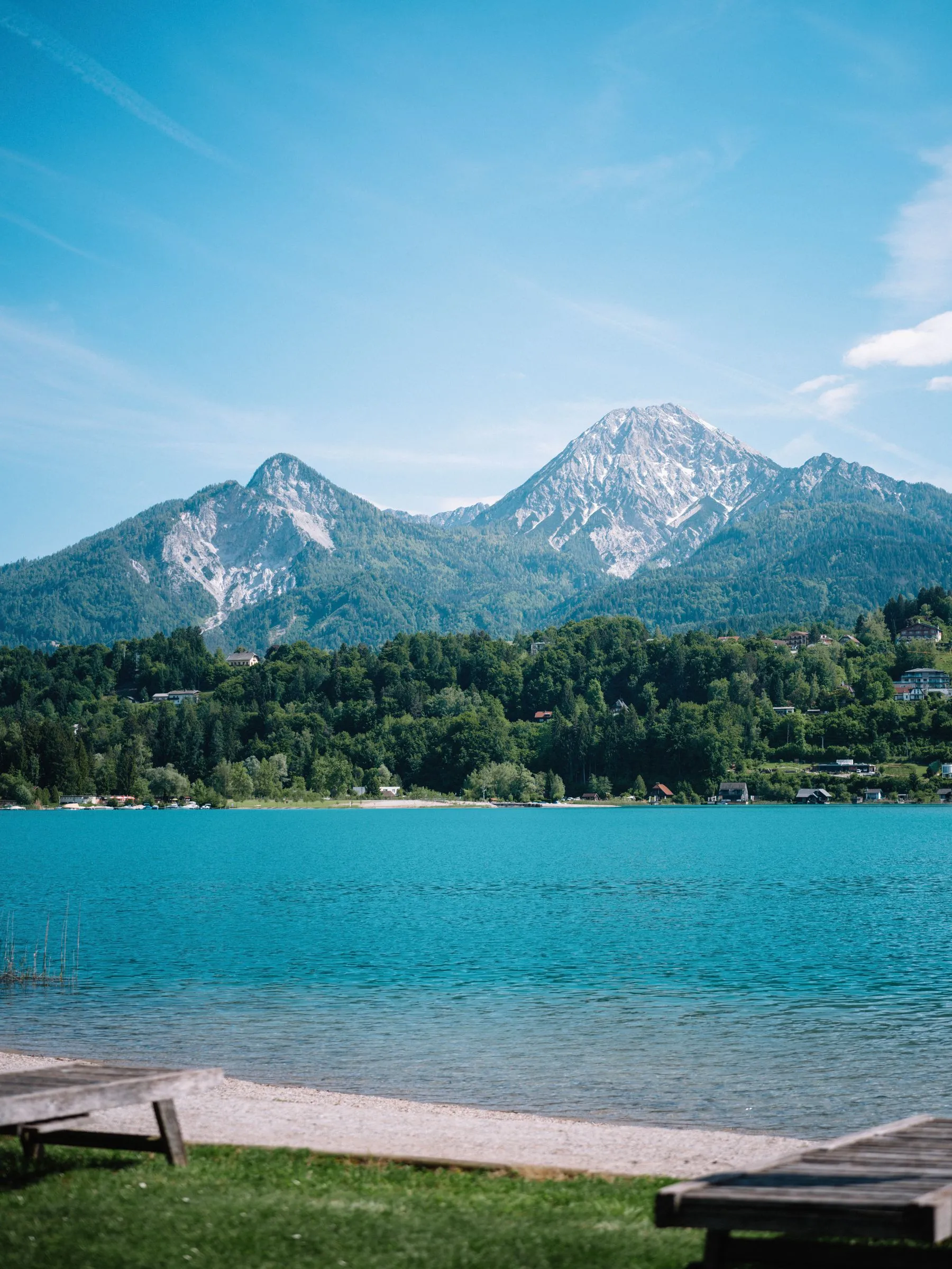 Der Badestrand mit Bäumen, dahinter der See und die Berge.
