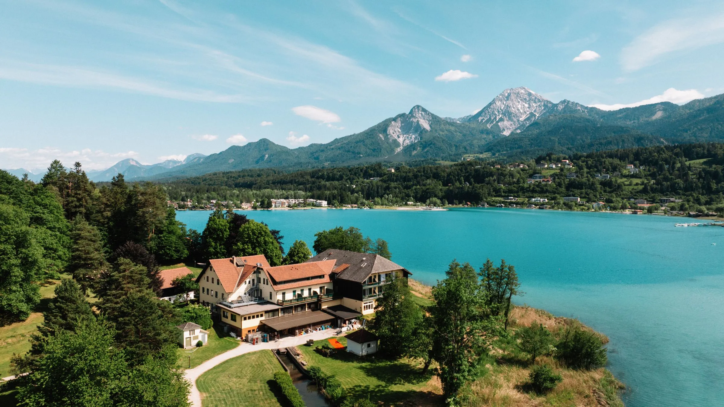Das Hotel auf der Insel, vom Wasser aus gesehen: Haupthaus, Bäume, dahinter die Berge.