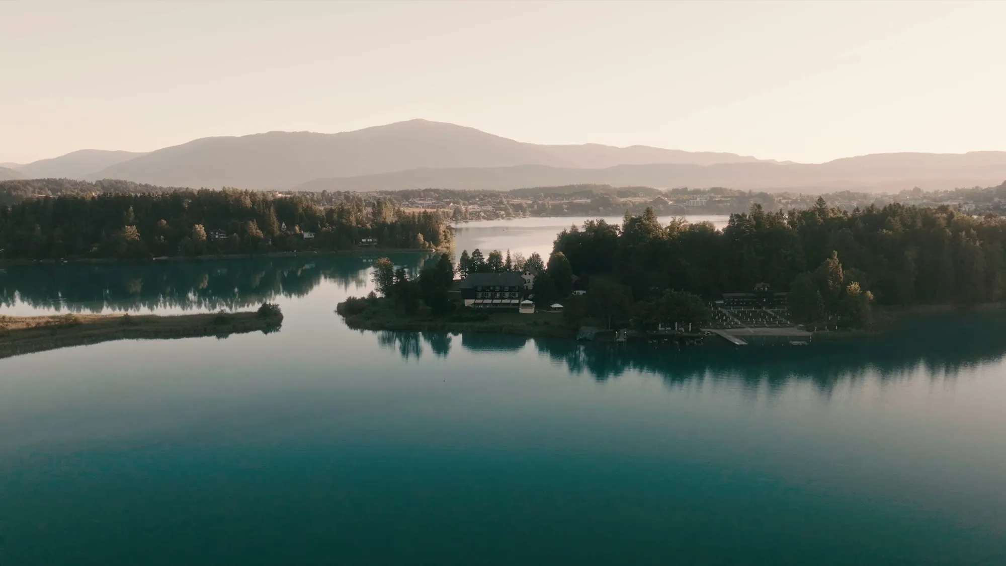 Die Insel im Morgenlicht, vom Wasser aus gesehen: das Haus zwischen den Bäumen, davor der stille See.