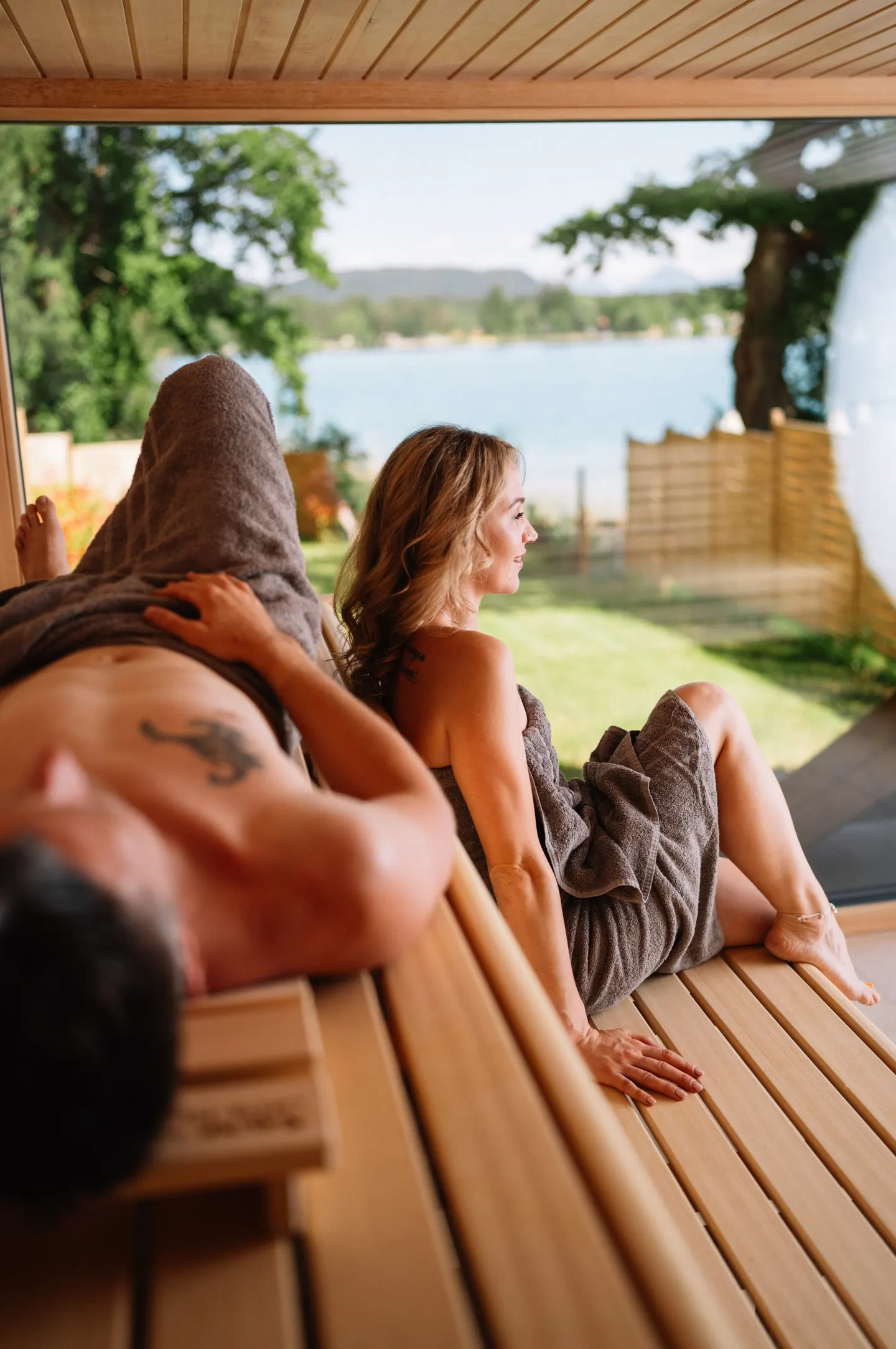 Zwei Personen ruhen in der Sauna vor dem großen Fenster zum Faaker See
